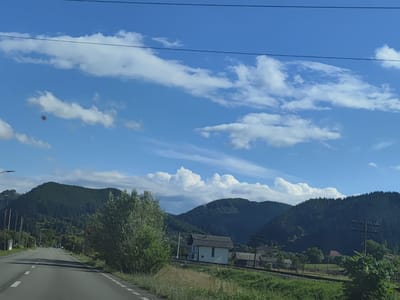 A view from a car on a paved road in Leordina, Romania, shows a rural landscape with mountains, trees, and a few buildings. The road curves to the left in the foreground, with white dashed lines marking the lanes. On the right side of the road, there is a grassy area with some trees and bushes. Further back, a white house with a dark roof is visible, along with other structures and a railway track. The background is dominated by green, tree-covered mountains under a bright blue sky with scattered white clouds. Overhead power lines are visible in the upper portion of the image. The lighting suggests it is daytime, likely midday.