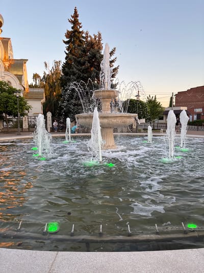 The image shows a multi-tiered fountain with several water jets spraying upwards, creating a frothy effect in the water. The fountain is illuminated with green lights from below the water's surface. In the background, a building with a golden dome and white walls is visible, suggesting a church or religious structure. Evergreen trees and other foliage are present, along with some urban infrastructure like buildings and a fence. A few people are seated on a bench in the middle ground, and one person is standing near the fountain in the background. The sky is clear and light blue, indicating daytime. The scene appears to be a public square or park area within the city of Tbilisskaya, Russia.
