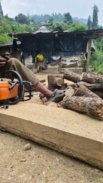 The image shows a person, partially visible, in an outdoor, rural setting. The person is wearing brown pants and a grey Croc-style shoe. Their arm is extended, and they are holding a grey sandal. In the foreground, an orange and black Stihl chainsaw rests on the ground. To the right of the chainsaw, a rough-hewn wooden plank lies horizontally. Behind the person and chainsaw, a pile of cut logs is visible, stacked against a makeshift structure. This structure appears to be made of wood and covered with black plastic sheeting and possibly blue tarpaulin. Further in the background, a lush, green hillside with scattered trees and vegetation dominates the landscape. The sky is overcast, suggesting a cloudy day. The overall scene conveys a sense of manual labor in a remote, mountainous area, possibly in Shila, India, given the context. The visual elements suggest a logging or woodworking activity.