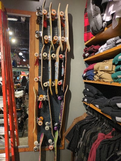 A collection of skateboards is mounted on a pegboard, displayed against a gray wall. Above, clothing hangs from wooden shelves. A mirror and what appears to be a ladder are visible below the skateboards. The arrangement creates a retail environment with an emphasis on skateboarding culture. The lighting is slightly dim, casting shadows that highlight the textures of the various items.