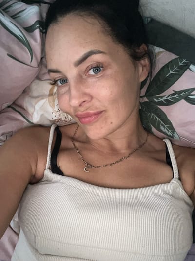 A close-up, selfie-style shot captures a woman lying down, her head and upper torso filling the frame. She has blue eyes, dark hair pulled back, and is wearing a white ribbed tank top and a delicate necklace with a crescent moon charm. The background is a pillow with a pink and green floral pattern. The image does not contain any information that would allow for the determination of its location in Maloye Isakovo, Russia.