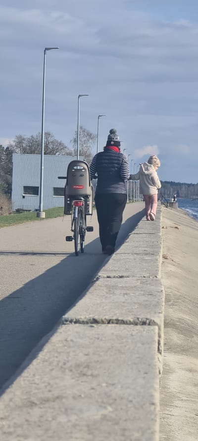 The image captures a bright, sunny daytime scene along a paved waterfront path in Ikšķile, Latvia. The perspective is slightly from behind and to the side of two individuals.

In the foreground and mid-ground, an adult, viewed from the back, is walking while pushing a bicycle equipped with a grey "POLISPORT" child seat. The adult wears a dark puffy vest over a striped long-sleeve shirt, dark pants, and a grey knit beanie featuring a red patch with partially visible white text that appears to say "RIDERS" or "RANDERS". Ahead of the adult, a young child or toddler stands, also viewed from behind, looking out towards the water. The child wears a light-colored hooded top, pink pants, and a patterned beanie.

The setting features a wide, grey paved path. To the right, a substantial concrete wall or barrier separates the path from a body of water, likely the Daugava River, which stretches into the background under a partly cloudy sky. On the left side of the path, several metal poles with L-shaped tops, resembling streetlights or supports, extend upwards. Behind these poles, a low, light grey corrugated metal building with two square windows is visible, flanked by some bare trees.

Long shadows cast across the path from the concrete barrier and the individuals suggest either a mid-morning or late afternoon time. The overall impression is one of a peaceful, leisurely outdoor stroll or bike ride along the riverbank on a clear day.