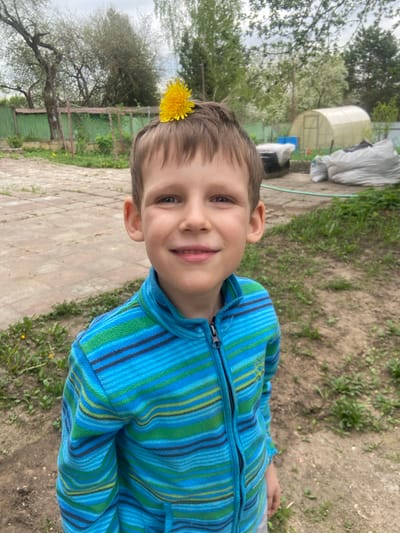 A boy with light brown hair is smiling at the camera with a dandelion flower perched on his head. He is wearing a blue and green striped fleece jacket. He is standing on a light-colored, paved surface, possibly a patio or walkway. Behind him is a grassy area with trees and a white greenhouse structure. The location is likely Verkhnee Myachkovo, Russia.
