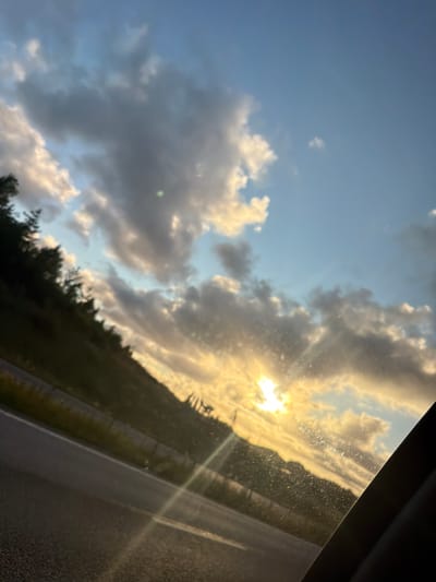 The image shows a view from inside a vehicle looking out at a sunset. The sky is a mix of blue and cloudy, with the sun's rays breaking through the clouds, creating a golden glow. There is a hillside on the left side of the frame, covered in trees, and a road in the foreground. The scene appears to be a rural or semi-rural area, possibly on the outskirts of Uddevalla, Sweden. There are no people or specific landmarks visible that would definitively pinpoint the location within Uddevalla.