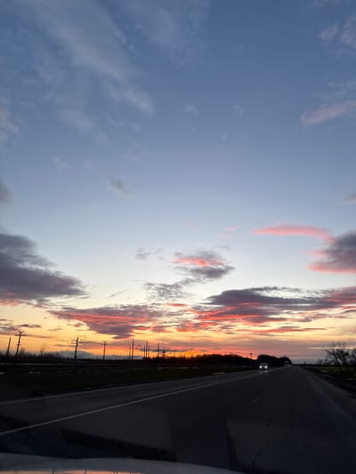 A sunset sky filled with a variety of clouds tinged with orange and pink hues stretches across a landscape. Utility poles line the horizon, creating a sense of depth. A road occupies the right side of the image, with two cars visible in the distance. The perspective suggests the photo was taken from inside a vehicle. The sky transitions from a pale blue to vibrant shades of orange and red closer to the horizon.