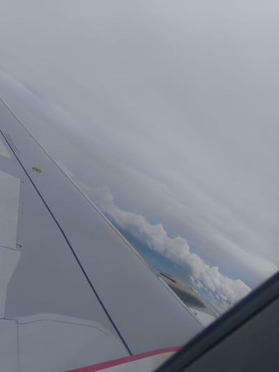 The image is taken from the window of an airplane, looking out over a wing. The wing is white with some gray lines and rivets visible. There is a small yellow object on the wing. Text is visible on the wing, but it is too blurry to read. In the distance, there are clouds and a blue sky. The time of day appears to be daytime, and the weather seems to be clear with some clouds. The setting is an airplane in flight. The image conveys a sense of travel and observation from above.