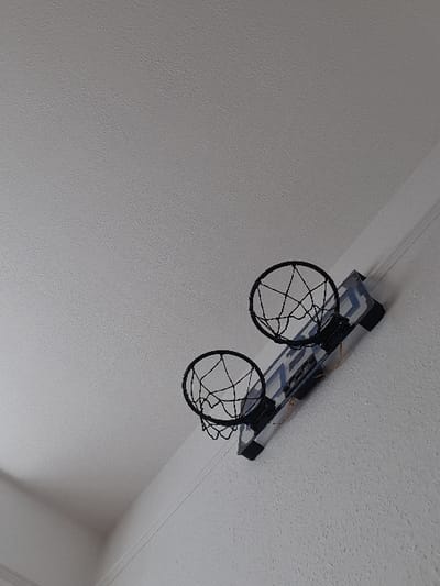 The scene is an interior room within Chatham, United Kingdom. The image presents a low-angle view looking upward towards a white, textured ceiling in the upper left and a matching textured white wall extending into the lower right. Diagonally mounted on the wall is a miniature dual-hoop basketball system. This system consists of a white rectangular backboard featuring blue graphic elements, below which are two black metal basketball hoops, each equipped with a black nylon net. Thin, orange-colored wires are visible underneath the lower edge of the backboard. No individuals or active interactions are present in the frame.