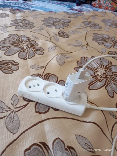 A white power strip with two outlets is plugged into a wall socket. A white phone charger is plugged into one of the outlets, and its white cable extends out of frame. The power strip and charger are on a beige fabric with a floral pattern. The image has a timestamp in the bottom right corner that reads "03.07.2026 14:42". There is no visible indication of the location within the city of Октябрь, Kyrgyzstan.