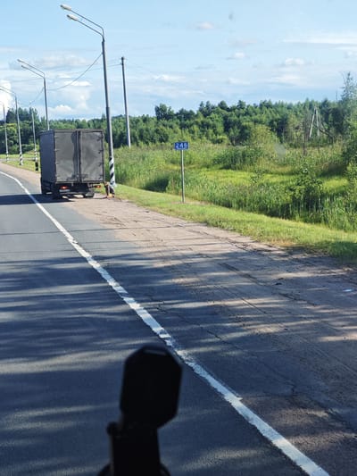A semi-trailer truck is parked on the shoulder of a road. A person in a dark shirt and shorts is kneeling beside the truck, appearing to be working on it or inspecting something on the ground. The truck is grey and has its rear doors closed. A blue sign with the number "246" is visible to the right of the truck, by the side of the road. The scene is set in a rural environment with tall green grass and trees lining the opposite side of the road. The sky is blue with scattered white clouds. Power lines are visible overhead, supported by poles. The road has a white line marking the lane. The foreground shows a portion of a dark object, possibly a camera or mount, obscuring the lower part of the image.