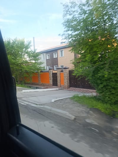 The image shows a street view in Svobodny, Russia, likely from inside a vehicle due to the presence of a car window frame in the foreground. A two-story building with a light beige facade and brick accents is visible behind a decorative fence. The fence has a section of bright orange and stone pillars leading to large, dark brown gates. Trees with lush green foliage are scattered around the property and along the street. The road in front appears to be paved and slightly worn, with a concrete curb. The sky is a clear, light blue with a few faint clouds. The overall impression is a residential area with well-maintained property.