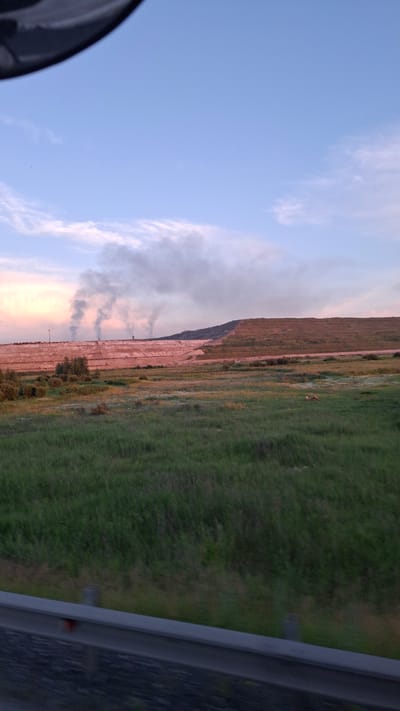 The image depicts a landscape scene with a clear sky, clouds, and a grassy field bordering a large embankment. The embankment appears to be part of a landfill or a construction site, with tiered layers and some vegetation growing on it. Smoke plumes are visible rising from the base of the embankment, suggesting an ongoing event such as a fire or industrial activity. In the distance, a few animals are present in the field. The sky displays a soft blend of blue and pink hues, indicating either sunrise or sunset. The foreground features a metallic railing, suggesting the viewer is observing the scene from an elevated platform or vehicle. There is no visible text in the image. The overall impression is one of a vast, somewhat desolate landscape experiencing an event that is generating smoke.