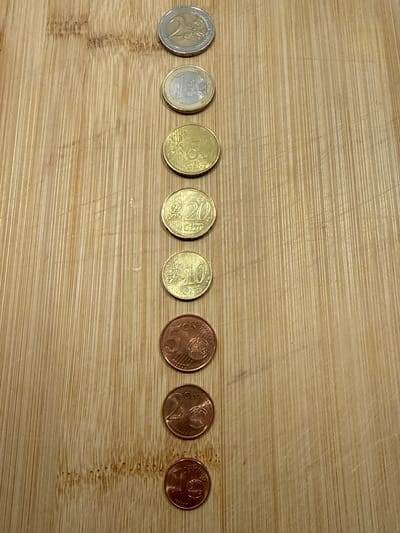 A total of eight euro coins are arranged vertically in a line on a wooden surface. The coins are of various denominations and metals, and appear to be from left to right: a 2 euro coin, a 1 euro coin, a 50 cent coin, a 20 cent coin, a 10 cent coin, a 5 cent coin, a 2 cent coin, and a 1 cent coin. The wooden surface is made of planks with visible grain and texture. There are no people or any other objects visible in the image. The scene is static and does not depict any action. This image was captured in Velbert, Germany.