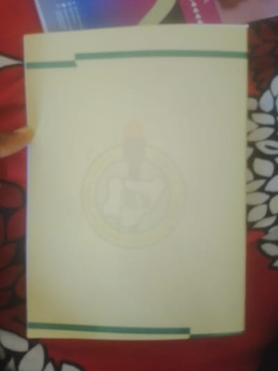 The image shows a pale yellow folder with a faded circular emblem in the center, featuring what appears to be a candle with flames. The folder has two horizontal green stripes near the top and bottom edges. It is placed on a red surface with a floral pattern. The image is blurry and poorly lit, making it difficult to discern further details of the emblem or the surrounding environment. There are no people or specific landmarks visible that would indicate a location within Magajin Gari, Nigeria, or any other city. The overall scene is a still life of an object, likely a document folder, on a patterned surface.