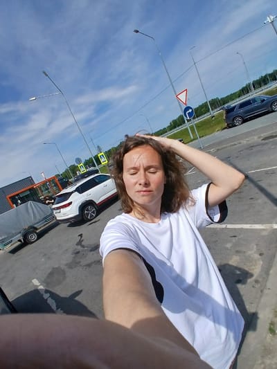 A medium-shot, selfie-style image captured outdoors during daylight. A woman with light brown hair is in the foreground, facing slightly away from the camera, with her right hand raised and resting on her head. She is wearing a white t-shirt with black trim on the sleeves. The background includes a parking lot with several cars, including a white SUV and a dark-colored sedan. A trailer is parked to the left of the white SUV. Traffic signs and streetlights are visible. The environment appears to be a roadside rest area or parking lot near a wooded area, consistent with a location in Akhmasikha, Russia, indicated by the presence of birch trees and the overall landscape. The sky is blue with scattered clouds.