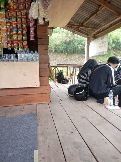 The image shows a wooden porch outside a small shop in Kecamatan Pangalengan, Indonesia (latitude: -7.233, longitude: 107.536). The shop's exterior is made of wood with a brown and orange color scheme. The porch is made of wooden planks and is slightly weathered. There is a gray rug laid out on the left side of the porch. Two people are sitting on the porch. The person on the left is wearing a dark gray sweater and black pants, while the person on the right is wearing a black jacket with a dark gray hood and black pants. The person on the right is facing the person on the left and is looking off into the distance. There are three black helmets sitting on the porch, one on each of the people and one by itself on the ground. The shop has a sign on the side facing the porch, displaying "DE LUKE GARAM". In the background, there are leafy green trees and a wooden railing. A rack of packaged snacks and bottles of water are displayed behind the counter inside the shop.