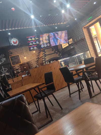 This media file depicts the interior of a cafe, likely in Petrovskaya, Russia, with a focus on a pizza and coffee menu. The scene is brightly lit with overhead lights and natural light streaming in from a large window or doorway on the right. The cafe has a rustic yet modern aesthetic, with exposed brick walls, wooden accents, and industrial-style furniture.

Prominently displayed is a digital menu board showing various pizza options, with titles like "ХАЧАПУРИ" and "СЕНДВИЧИ," along with prices. To the left, a vintage-style clock with a chef's hat graphic reads "ВКУСНО БЫСТРО" (Deliciously Fast). A smaller chalkboard menu lists coffee drinks such as "ЭС Преско," "АМЕРИКАНО," "КАПУЧИНО," "ЛАТТЕ," and "ЧАЙ," with their respective prices.

Several tables and chairs are visible, some empty and some occupied by what appear to be patrons. The chairs are upholstered in dark material, and the tables are wooden with metal frames. In the background, a counter area features glass display cases, possibly holding food items, and shelving with jars. A prominent "ВЫХОД" (Exit) sign is illuminated green above the doorway on the right. The overall impression is that of a casual and inviting dining establishment. The time of day appears to be daytime, given the natural light.