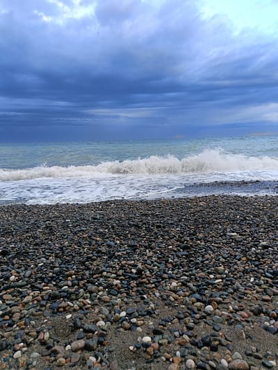 The image shows a pebble beach with waves crashing on the shore under a cloudy sky. The pebbles in the foreground are mostly dark in color, with variations of gray, black, and brown. The waves are white and foamy as they break. The sky is overcast with dark, heavy clouds. This is most likely the location in the city of Gulrypsh, Abkhazia. There are no people in the image.