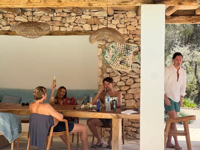 Four individuals are gathered around a rustic wooden dining table on a covered patio. The patio features a stone wall and exposed wooden beams. The people are dressed casually, suggesting a relaxed outdoor setting.
On the left, a shirtless man with blonde hair is seated, with a grey t-shirt draped over his shoulders. He faces away from the camera, looking towards the table. Next to him, a woman with brown hair is seated and holding up a bottle of beer. She is smiling and appears animated. She is wearing a patterned dress.
In the center, a man with brown hair is seated at the table, appearing to be on his phone. He is shirtless and wearing swim trunks. Various items are on the table in front of him, including bottles of water, a cooler, and what appear to be toiletries.
To the right of the patio, a man in a white shirt and green patterned swim trunks is standing, looking downwards. He is partially obscured by a white pillar.
The environment suggests a warm climate, with lush greenery visible in the background. The overall impression is one of a casual gathering or meal on a sunny day in a Mediterranean setting, possibly at a villa or a restaurant in Santa Eulària des Riu, Spain, given the architectural style and natural surroundings.