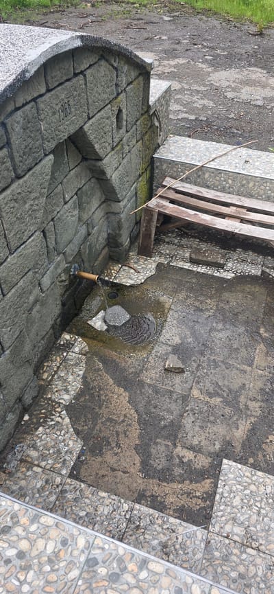 The image shows a stone water fountain in Polyanovo, Bulgaria, built in 1965. The fountain is made of large gray stone blocks. Water flows from a brown pipe into a small pool. A wooden pallet leans against the fountain. The surrounding area is a mix of stone tiles, bare ground, and patches of grass. The ground is wet and stained.