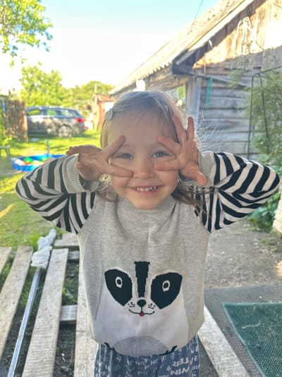 A young girl is standing outdoors, likely in the city of Piriutino, Russia. She is facing the camera with a playful expression, holding up her hands to her eyes in a gesture that resembles a peace sign. The girl is wearing a grey sweater with a raccoon design and striped sleeves, and patterned pants. Behind her, a wooden structure, possibly a shed or house, and a car are visible. The environment appears to be a yard or outdoor area, with trees and a sandbox in the background. The lighting suggests it is daytime.
