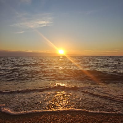 The image depicts a serene seascape at sunset. The sun, a bright, luminous orb, is positioned slightly left of center on the horizon, casting a warm, golden glow across the water and sky. Sunbeams radiate outwards from the sun, creating streaks of light that reflect brilliantly on the rippling surface of the sea. Small, gentle waves are breaking on a pebbly shore in the foreground, with frothy white foam tracing the edge of the water. In the distance, along the horizon line, faint structures resembling poles or buoys are visible. The sky transitions from a pale blue at the top to a warm, hazy orange near the horizon, with wispy clouds scattered across its expanse. The overall impression is one of tranquility and natural beauty during the golden hour. The location is identified as Chelyadinovske rural settlement, Ukraine.