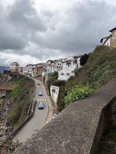The image depicts a steep coastal hillside in Colunga, Spain, under an overcast sky dominated by heavy grey clouds. A narrow asphalt road curves through the scene, descending from the upper left towards the lower center, then continuing further left out of frame. Three sedans are visible on the road, all traveling away from the viewer: a dark grey vehicle in the foreground on the right lane, a light blue vehicle further up on the left lane, and another dark grey vehicle higher up on the right lane. Orange traffic cones and yellow rectangular markings delineate sections of the road's right edge, which is bordered by a stone path. On the left side of the road, a concrete staircase with a white lamppost descends towards a rocky shoreline. Overhead wires traverse the area above this staircase.

The hillside to the left of the road drops sharply to the rocky coast, featuring abundant green and brown vegetation, with remnants of older stone structures and walls. To the right of the road, the hillside ascends steeply, covered in dense green foliage. Numerous multi-story buildings, characteristic of a coastal town, are clustered along the ascending road and further up the hillside. These structures exhibit varying architectural styles and colors, including white, cream, pink, yellow, and brown facades, some with visible wear and tear. A dark grey, textured concrete wall or barrier occupies the immediate lower right foreground, running diagonally across the corner. No individuals are visible.
