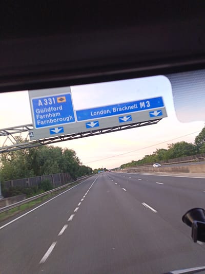 A highway sign is visible above a road. The sign is blue and white and displays route numbers and destinations. On the left, it shows "A 331" with "Guildford Farnham Farnborough" listed below. On the right, it shows "London, Bracknell M3". Two blue downward arrows are visible below each section of text, indicating directions. The road itself appears to be a multi-lane highway with white dashed lines marking the lanes. There is green foliage and trees on the sides of the road, and a wooden fence can be seen on the left. The sky is a pale pinkish hue, suggesting either sunrise or sunset. A white car is visible in the distance on the right side of the highway. The perspective suggests the image was taken from inside a vehicle.