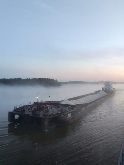 In the early morning, a towboat navigates a foggy river with a barge carrying a load of light-colored aggregate. The barge, marked with the number 7903, is positioned in the foreground and midground, its deck equipped with railings and various machinery. A single light illuminates part of the barge's deck. In the background, a dense line of trees borders the river, partially obscured by the morning mist. The sky above is a pale blue, transitioning to a warmer hue near the horizon, suggesting the rising sun. The river's surface is calm, with subtle ripples reflecting the muted light. The scene is devoid of human presence, focusing solely on the industrial vessel and the natural environment. The specific location in Chaika, Russia is not discernible from the image.