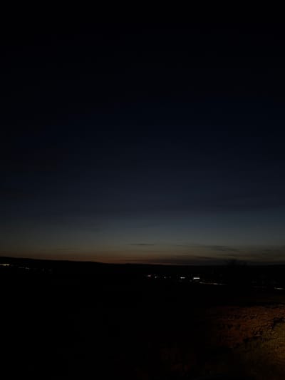 The image displays a panoramic, wide-angle view of the landscape at twilight or night, situated in the city of Ober-Mörlen, Germany. The upper two-thirds of the frame consist of a dark blue-grey sky, exhibiting a subtle gradient to a faint orange-brown hue along the horizon line. A slender crescent moon is discernible in the upper left quadrant of the sky, with a single bright, unidentifiable celestial body (star or planet) positioned slightly higher and to its right. Wispy, diffuse cloud formations are present in the sky.

The middle ground features a dark, undulating silhouette of hills or elevated terrain, forming a clear horizon line. Below this horizon, a collection of static artificial lights indicates the presence of the municipality of Ober-Mörlen, with distinct clusters suggesting buildings or street illumination. The foreground, occupying the lower third of the image, is largely obscured by darkness, rendering specific objects indistinguishable. A faint, reddish-brown patch of ground, possibly dirt or sparse vegetation, is subtly visible in the lower right corner, indicating the immediate terrain. No human figures, animals, or active elements are depicted.