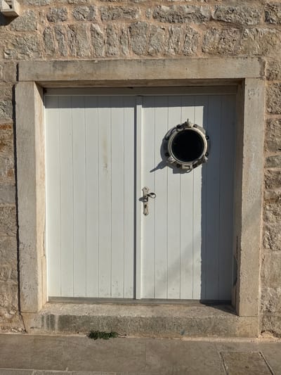 The image shows a weathered, white wooden double door set within a stone facade. The door is framed by a rustic stone architrave and sits on a stone threshold. A round, metal porthole window is affixed to the right-hand door. The stone wall surrounding the door is constructed from rough-hewn blocks. The ground in front of the door is paved with stone. The overall impression is one of age and solidity, suggesting a location with a long history, possibly in a coastal area. The presence of the porthole window hints at a maritime connection, or perhaps an architectural style that evokes such a theme. The lighting suggests it is daytime, with distinct shadows cast by the door and the porthole. The location is identified as Jelsa, Croatia.