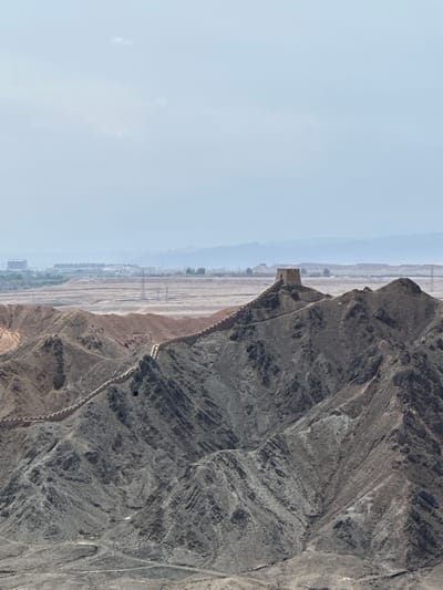 The image depicts a panoramic view of the Jiayu Pass fortress, also known as the Great Wall of China, in Jiayuguan, China. The fortress is situated on a rocky, mountainous terrain, with a long, winding wall extending across the landscape. A prominent watchtower stands atop a peak, overlooking the surrounding area. In the distance, a hazy cityscape is visible, with buildings and infrastructure indicative of a developed area. The sky is overcast and pale, with a few wispy clouds. The overall impression is one of historical significance and rugged natural beauty.