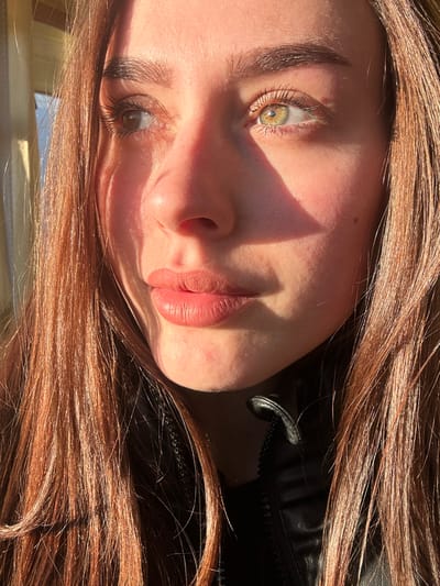 A close-up, cropped portrait captures the face of an individual, specified as being located in Muirhead, United Kingdom. The subject, a female with fair skin and long, reddish-brown hair, is illuminated by strong, warm, directional sunlight originating from the viewer's left. Her left eye (viewer's left) displays a hazel-brown color, while her right eye (viewer's right) exhibits a green-hazel hue, both gazing slightly upward and to the left. Pronounced shadows are cast across the right side of her nose, beneath her right cheekbone, and along the right side of her face. Her lips are closed, presenting a soft pink, matte appearance. A small, dark mole is visible on her right cheek. The subject is wearing a black collared jacket, possibly leather, with a partially open zipper visible at the neckline. The background is brightly lit and out of focus, suggesting an indoor setting near a light source or window.