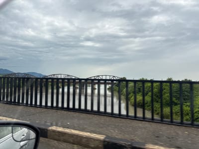 The image shows a view from a bridge over a river in Samtredia, Georgia. The sky is overcast with clouds. In the distance, two railway bridges with red trusses span the muddy river. The riverbanks are lined with lush green trees. A black metal railing runs along the edge of the bridge in the foreground. Part of a car's side mirror is visible on the left side of the image, indicating the viewpoint is from a moving vehicle. The road surface of the bridge is visible, appearing wet and reflective.