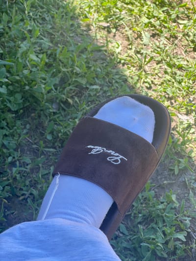 The image shows a close-up, high-angle shot of a person's left foot wearing a light blue sock and a brown slide sandal. The sandal has white embroidered lettering on its strap, which appears to be a stylized logo or brand name. The foot is resting on a patch of green grass with dry soil visible beneath. The background consists of more grass and soil, suggesting an outdoor environment, possibly a park or yard. The lighting indicates it is daytime, with shadows cast by the foot and sandal. No specific location in Zudilovo, Russia, can be determined from the visual information provided.
