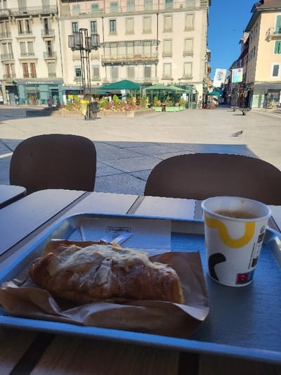 An outdoor cafe scene captured on a sunny day in Place des Arts, Thonon-les-Bains, France. In the foreground, a light-grey serving tray rests on a dark-slatted table, featuring a single golden-brown croissant topped with a pale, possibly almond, crust, situated on brown paper. Alongside the croissant is a white paper cup with a yellow and black abstract design, containing a light-brown beverage, likely coffee, topped with crema. Two dark-brown, rounded chair backs partially obscure the lower view.
The midground reveals a spacious, light-paved public square under bright sunlight, casting distinct shadows. A multi-branched modern streetlamp stands to the left of center. A large, raised planter filled with green foliage and colorful flowers occupies the center. Behind the planter, two green parasols shade an outdoor cafe area with tables and chairs. Two individuals are visible walking to the left of the planter, and a pigeon is on the pavement to the right.
The background is dominated by multi-story European architecture. To the left and center, a large, light-colored building with numerous windows and light-colored shutters defines the square's edge, hosting ground-floor businesses, including one with a green awning and another with blue signage. To the right, a yellowish building extends down a street, also featuring windows with green shutters. The sky is clear and blue.