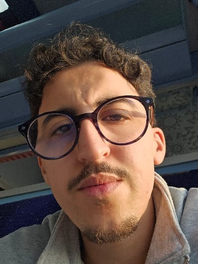 A close-up image depicts a young adult male in what appears to be an indoor setting. The male has light-to-medium skin, dark curly hair, and wears dark-rimmed round spectacles. He has a mustache and a short goatee on his chin. His expression is neutral, with a direct gaze and faint furrows visible on his forehead. He is wearing a light grey hooded garment, visible around his neck and shoulder. The background is out of focus, showing a dark overhead beam-like structure in the upper left, a light-colored patterned surface in the upper right, and a dark blue speckled surface (possibly fabric upholstery) in the lower left. The scene is located in Jbilate الجبيلات, Morocco.