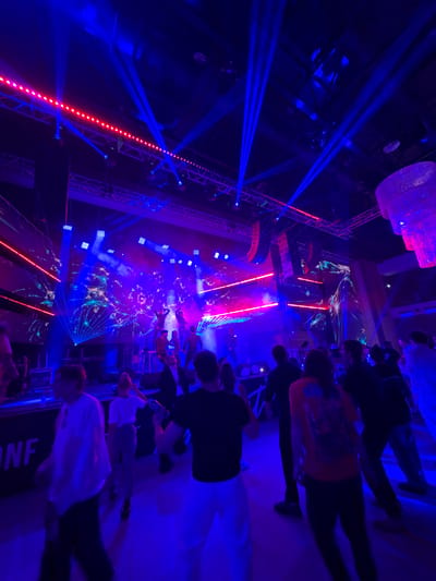 This is a photograph of a concert or performance taking place in an indoor venue in Tsilkani, Georgia. The scene is dominated by vibrant blue and pink stage lighting, with numerous laser beams projected across the space. In the background, a stage is set up with performers visible. The stage features large screens displaying abstract, colorful graphics. Above the stage, lighting rigs and speakers are suspended from the ceiling. In the foreground and midground, a crowd of people are present, some appearing to be dancing and others standing and watching the performance. The overall atmosphere suggests a lively event.