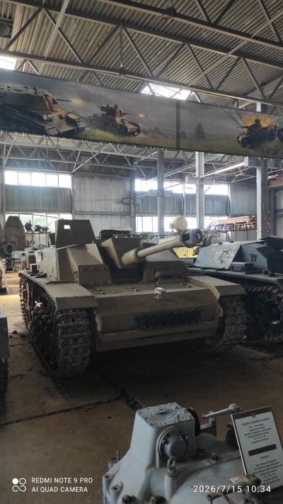 In an indoor exhibition space, likely a museum, a tan-colored armored vehicle, identified as a Sturmgeschütz III (StuG III) assault gun, is prominently displayed. The vehicle is positioned centrally, facing the viewer, with its tracks resting on a concrete floor. The StuG III features a casemate superstructure and a long gun barrel extending forward. In the background, other military vehicles and equipment are visible, some partially obscured. A large mural depicting tanks in action stretches across the upper portion of the scene. The environment appears to be a large industrial or hangar-like building with a metal roof structure and windows allowing natural light to enter. The image is dated July 15, 2026, and includes a watermark indicating it was taken with a "REDMI NOTE 9 PRO AI QUAD CAMERA." A sign in Russian and English, describing a gearbox, is partially visible in the foreground. This scene is located in the vicinity of Лашанскі сельскі Савет, Belarus, suggesting a military museum or historical site within that region.