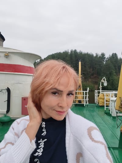 A woman with short, light peach-colored hair is taking a selfie on the deck of a ferry. She is wearing a white knitted cardigan with a beige zig-zag pattern over a dark blue t-shirt with white script. Her right hand is touching her hair. The ferry deck is green and wet, with some metal railings and machinery visible. In the background, a wooded shoreline is visible under an overcast sky. The image appears to be taken in Goritsy, Russia, likely near the Kirillo-Belozersky Monastery, which is accessible by ferry.