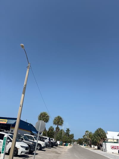 The image depicts a street scene in Holmes Beach, Florida, United States of America, on a clear, sunny day with a bright blue sky. In the foreground, a concrete utility pole stands to the left, supporting electrical wires. Adjacent to the pole is a white car with a green and white sign that reads "MARINA DR". Behind the car, a line of parked vehicles, including SUVs and cars, are visible. To the left, a building with a blue facade and yellow lettering that reads "AUTOMOTIVE" is partially visible. Further down the street, palm trees and other vegetation line both sides. In the distance, the street recedes towards a row of buildings. On the right side of the street, a white PODS container with "Moving & Storage" branding is parked. The environment appears to be a commercial or mixed-use area, possibly near a coastal location, given the presence of palm trees and the name of the street. There are no people clearly visible in the image.