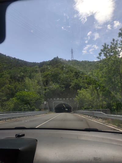 The image depicts a view from inside a vehicle driving towards a tunnel. The tunnel is flanked by a hillside covered in green trees. The entrance to the tunnel is decorated with a black and white checkered pattern. To the left, a utility pole stands near the road, which is separated from the hillside by metal guardrails. The sky is blue with scattered clouds, and power lines stretch across the upper part of the frame. The dashboard of the vehicle is visible in the foreground, slightly obscuring the bottom of the scene. Based on the presence of the tunnel, hillside, and general geography, this image is likely of the tunnel in Jeleznitsa, Bulgaria.