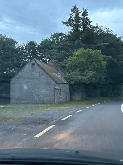 The image depicts a rural roadside scene in Rathvilly, Ireland, under an overcast sky. A grey stone building with a moss-covered, sloping roof stands to the left of a paved road. The building has a small, square window in its gable and a larger rectangular opening on its side. Dense green trees and foliage surround the building and extend behind it. The road, marked with yellow dashed lines, curves to the right and occupies the foreground. The perspective suggests the photo was taken from a moving vehicle.