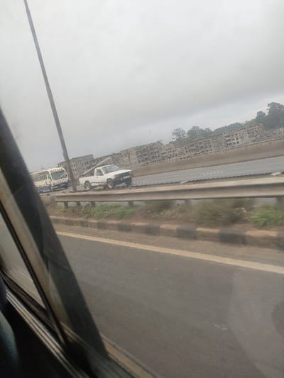The image depicts an outdoor scene from the perspective of a moving vehicle, looking across multiple highway lanes under an overcast sky in Kahawa Sukari ward, Kenya. In the immediate foreground on the left, a portion of the viewer's vehicle window frame is visible, with a hint of blue fabric. The lower section of the frame shows asphalt road with a solid white line bordering a raised curb.
Across the midground, a silver metal guardrail runs horizontally. Behind this barrier, a strip of reddish-brown earth with sparse green vegetation is present, followed by a black and tan painted curb. Beyond this, on additional highway lanes, a white pickup truck with visible front-end damage and dark wheels is depicted moving from left to right. Behind the truck, partially obscured by a dark utility pole on the far left, a white and blue Nissan passenger bus (matatu) is also moving in the same direction. Dashed white lines mark the highway lanes.
The background features a long line of multi-story concrete apartment buildings, many appearing to be in various stages of construction or unfinished, stretching across the horizon. To the far right, a cluster of green trees is visible on an elevated terrain. The sky is uniformly light gray and overcast, covering the entire upper half of the image.