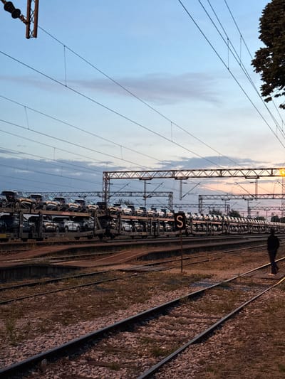 The image depicts a railway scene in Ruma Municipality, Serbia. A train loaded with cars is stationary on the tracks. Overhead electrical lines and support structures are visible. The sky is partially cloudy, suggesting either dusk or dawn. The railway tracks are surrounded by gravel and vegetation. The scene conveys an industrial or transportation hub environment. A sign with the letter "S" is visible along the train. There is a shadow of a person in the upper right corner of the frame.