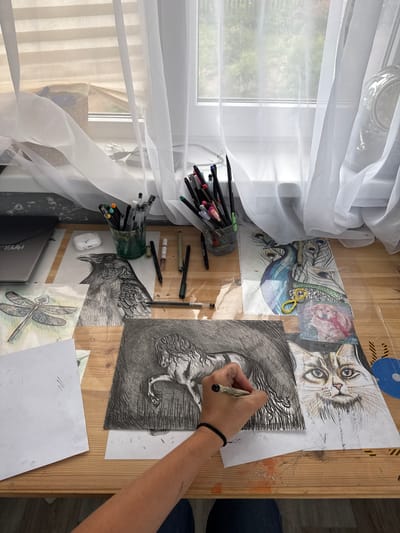 The image shows an artist's desk in Oliki, Russia, with several drawings and art supplies. The desk is made of wood and is covered with papers featuring drawings of animals, including a raven, a dragonfly, a peacock, a cat, and a horse. The artist's hand is visible, holding a pen and working on the drawing of the horse. Various pens and pencils are arranged in a glass container. A laptop and a wireless earbud case are also on the desk. A window with white curtains is in the background, allowing natural light to illuminate the workspace.