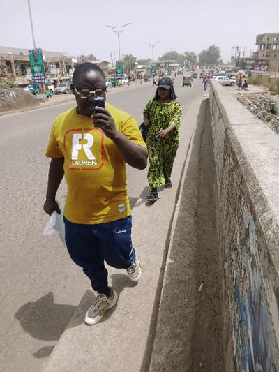 The image depicts a daytime street scene in Kadoma, Zimbabwe, under a bright, hazy sky. In the foreground, a male individual walks on a concrete sidewalk, holding a dark smartphone to his right ear and a folded white item in his left hand. He wears a mustard yellow t-shirt featuring a red 'R' with "LAUREN" text, dark blue track pants, light-colored sneakers, and eyeglasses. Following him, a female individual walks on the same path, dressed in a long green and black patterned dress, a dark baseball cap, and dark sandals. She carries a dark shoulder bag on her left side and a small light-colored object in her right hand.

To the left of the pedestrians, a paved asphalt road extends into the background, accommodating vehicular traffic, including cars and several green auto-rickshaws. The left side of the road is lined with commercial buildings displaying "Opay" advertising banners on street light poles. A concrete barrier runs along the right edge of the sidewalk, separating it from an unkempt area containing debris and sparse vegetation. In the far background on the right, a multi-story structure, possibly under construction or in disrepair, is visible. Shadows cast by the subjects indicate direct overhead sunlight.
