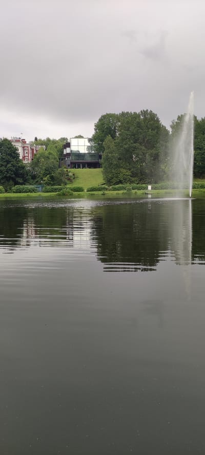 The image depicts a serene lake scene with a modern glass-fronted building and a fountain on the far bank. To the left, a more traditional red-brick building is visible, also nestled amongst lush greenery. The sky is overcast, casting a soft, diffused light over the landscape. The water of the lake is calm, with gentle ripples reflecting the surrounding trees and buildings. The overall impression is one of tranquility and understated elegance. This scene is located in Angelovo, Russia.