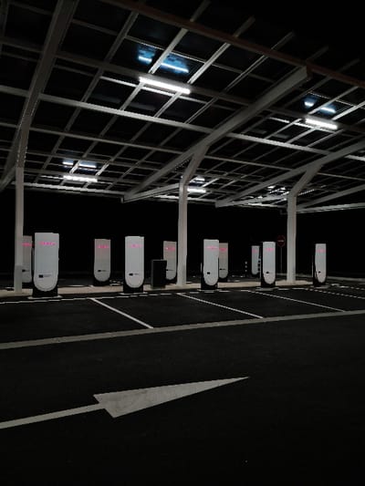 The image displays an unoccupied electric vehicle (EV) charging station at night in Les Tourrettes, France. A large, grey metal canopy structure, featuring a grid framework supporting dark rectangular panels (likely solar) and illuminated by multiple white fluorescent lights, covers the charging area. Nine modern, white charging pedestals with black lower sections are visible, each displaying a glowing pinkish-red light strip near its top. The ground is dark asphalt with white parking space lines and a large white directional arrow in the foreground, pointing towards the pedestals. A dark utility box and a red circular 'no entry' sign on a pole are also present. No individuals or vehicles are depicted in the scene.