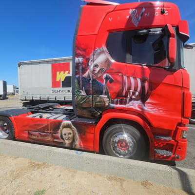 In Vinassan, France, a bright red truck is parked on a dusty roadside next to a concrete curb. The truck features elaborate airbrushed artwork. On the side of the cab, a Viking warrior with a beard and axe is depicted. Below this, on the lower body of the truck, a woman with blonde hair and a Viking ship are painted. The truck's side also displays the text "Lupo-Art AIRBRUSH." In the background, a white trailer with a red and yellow logo and the word "SERVICIO" is visible. The sky is clear and blue.