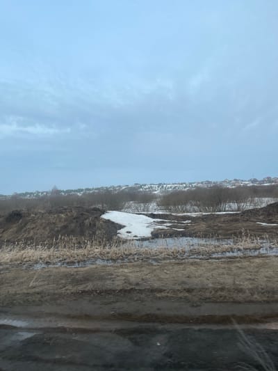 The image depicts a landscape with a focus on the transition from late winter to early spring. A patch of melting snow is visible in the middle ground, contrasting with the surrounding brown, dormant vegetation. In the background, a row of houses is visible, indicating a residential area. The overcast sky suggests a cold and potentially damp atmosphere. The right side of the image is blurred and distorted, likely due to being taken through a window or reflective surface, which obscures the full details of the foreground. Given the prompt's location of Yus'ki, Russia, the landscape is likely representative of the environmental conditions typically found in that region during the transition between seasons. There are no people visible in the frame. The primary subjects are the natural elements (snow, vegetation, sky) and the built environment (houses). The main "action" is the thawing process of the snow.